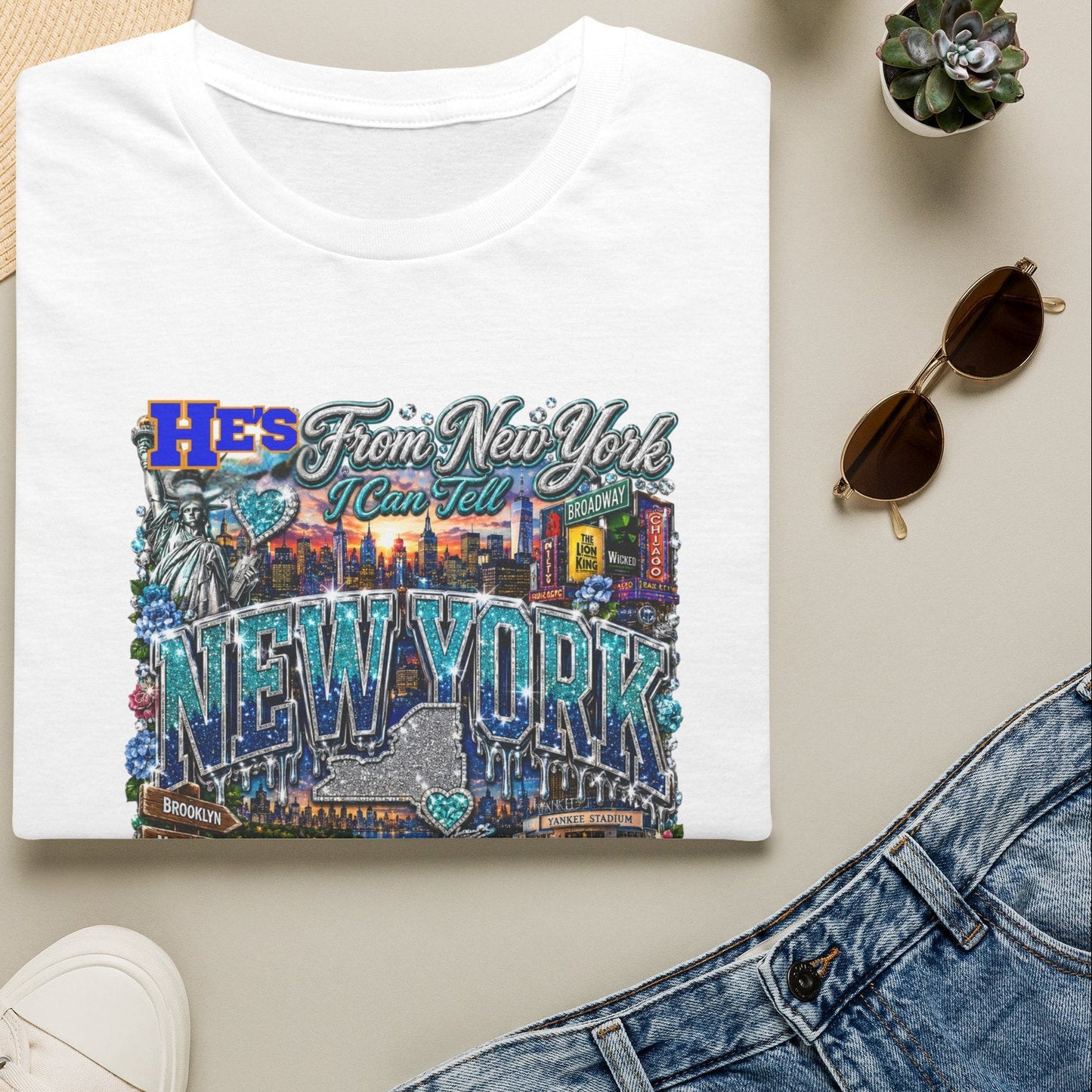 White t-shirt with 'He's From New York' design, surrounded by a hat, sunglasses, shoes, and plants on a beige surface.
